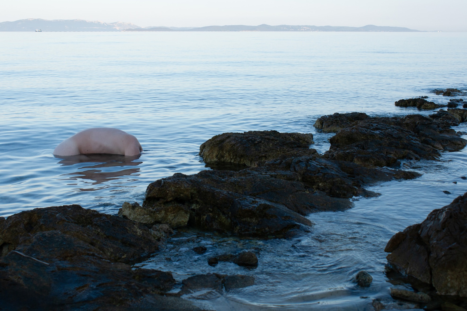 A figure partly submerged in the sea beside a rocky coastline