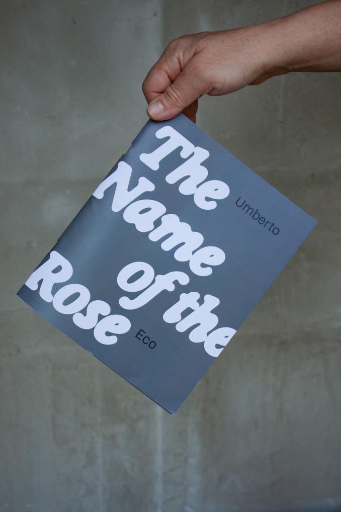 A hand holding a blue publication titled The Name of the Rose